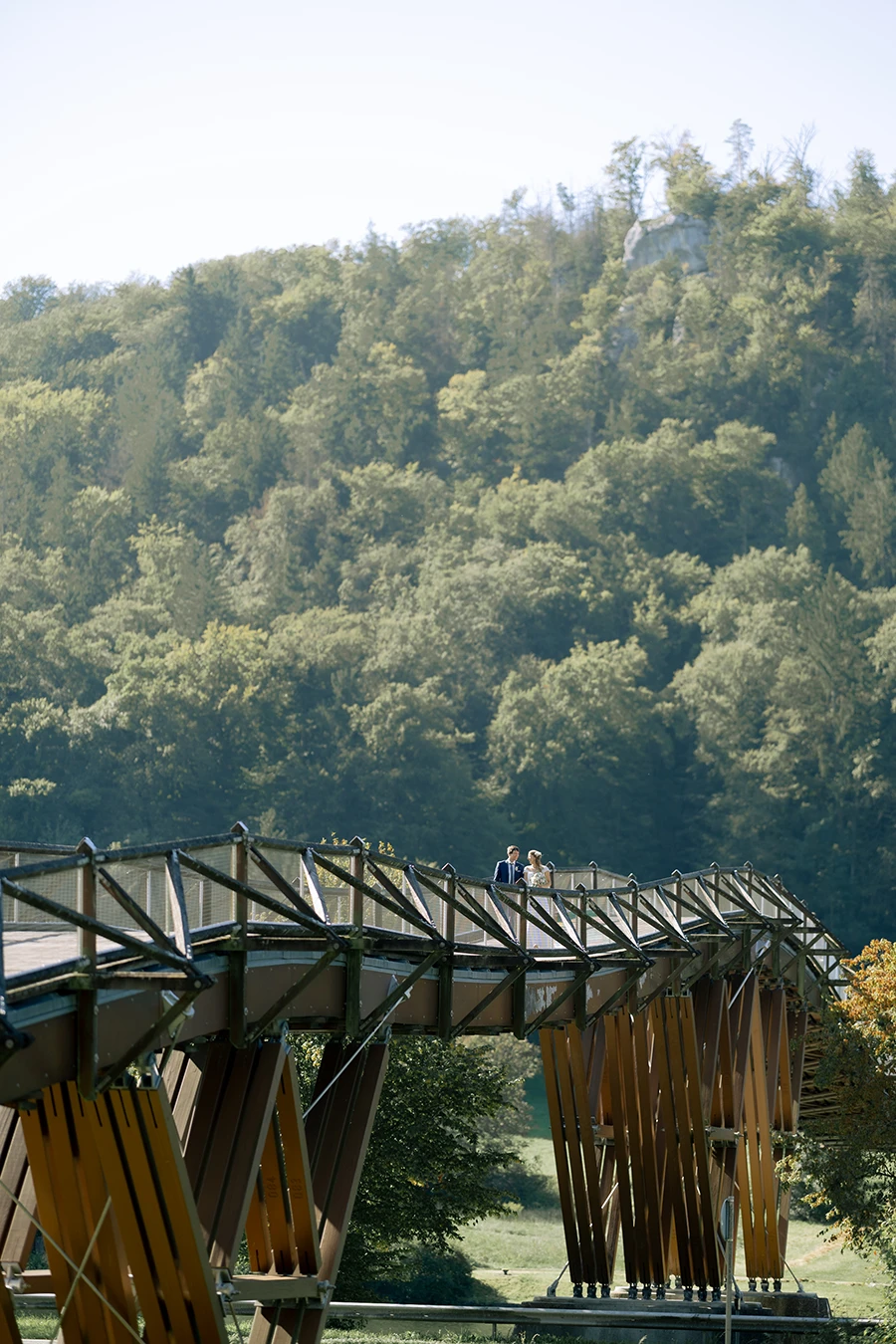 Hochkantaufnahme einer Holzbrücke über einen Fluss, klein kann man ein Brautpaar erkennen.